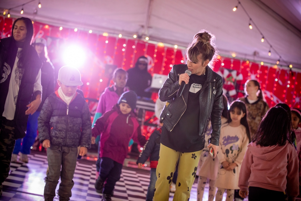 Woman holding microphone leads a group of children in a dance